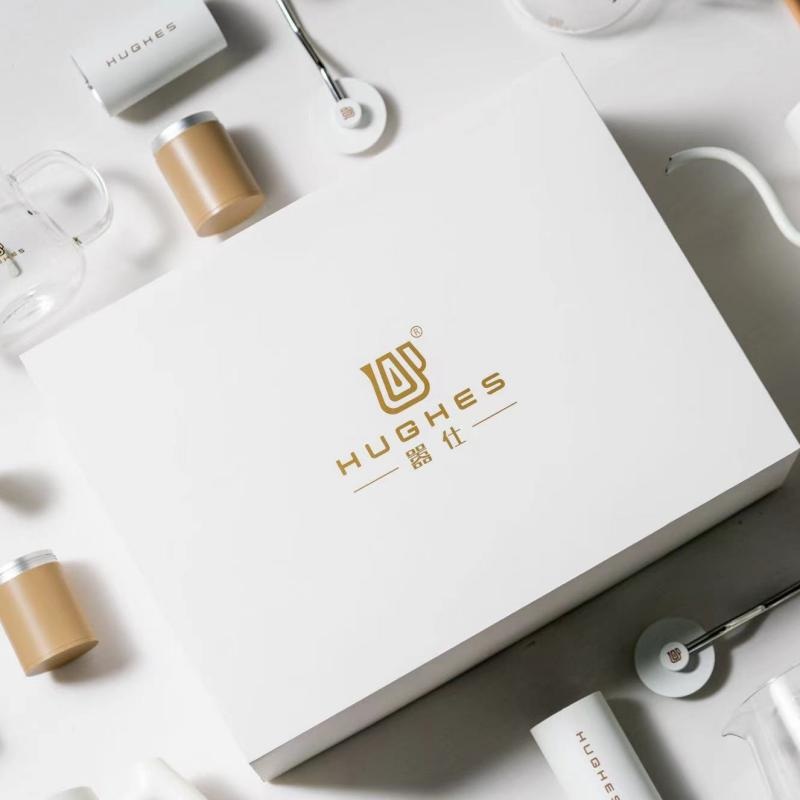 White rigid coffee gift box with gold foil HUGHES logo, flat lay with stainless canisters, a glass brewer and a wooden-handled cleaning brush